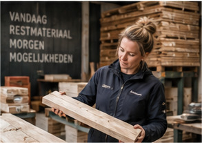 Vrouw in werkkleding tilt een houten balk uit een stapel in een werkplaats, met achter haar een bord met de tekst vandaag restmateriaal, morgen mogelijkheden.